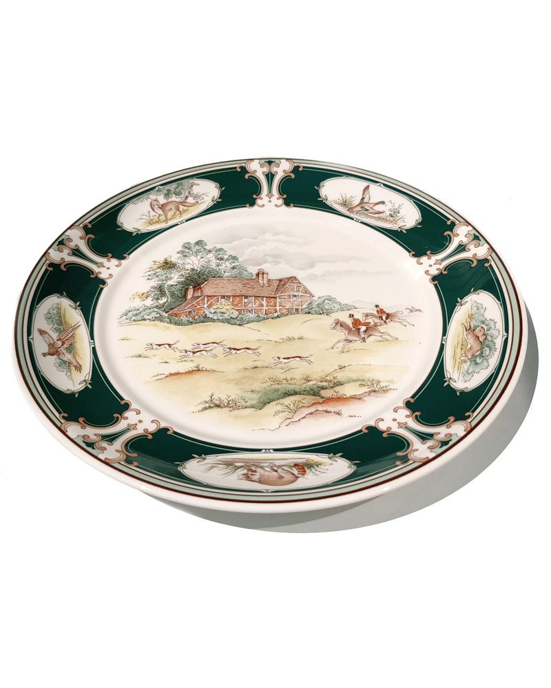 A decorative ceramic plate featuring a hunting landscape scene in the center, surrounded by a dark green border with gold detailing and small oval illustrations.